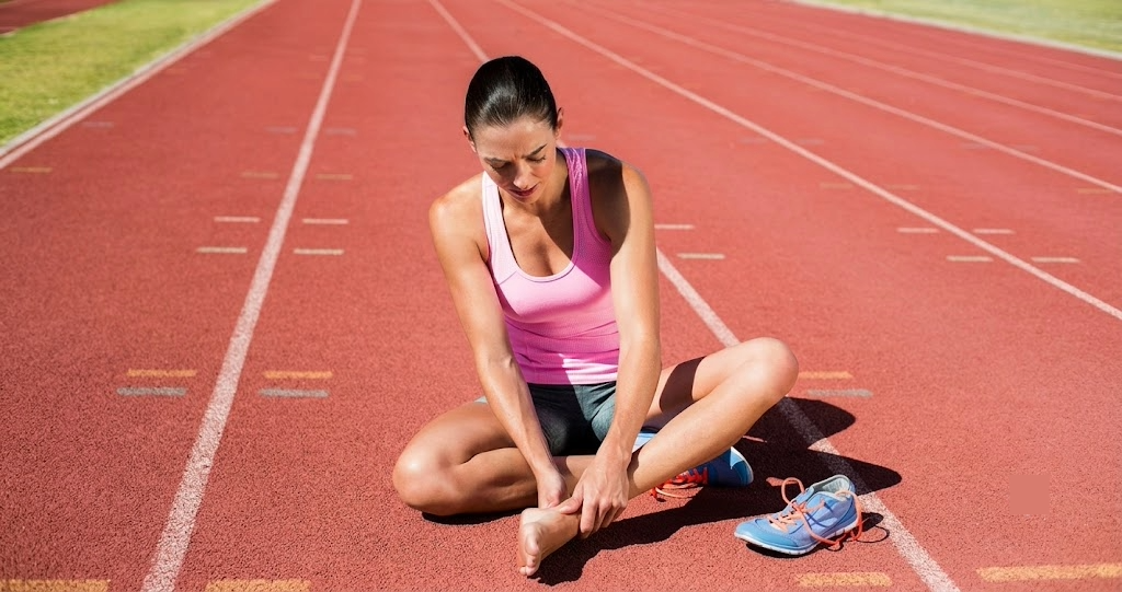 A female runner sitting on a track holding her painful bare foot and ankle after removing her running shoe, illustrating foot pain during exercise.